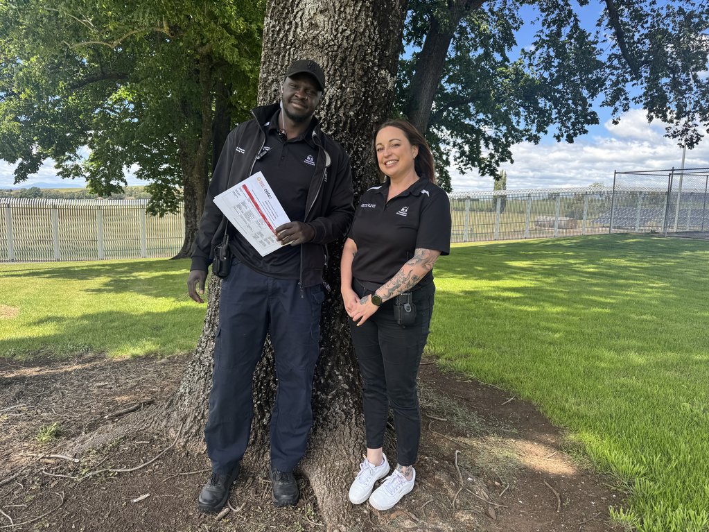 Two people wearing black leaning against a tree in a yard with a fence in the background